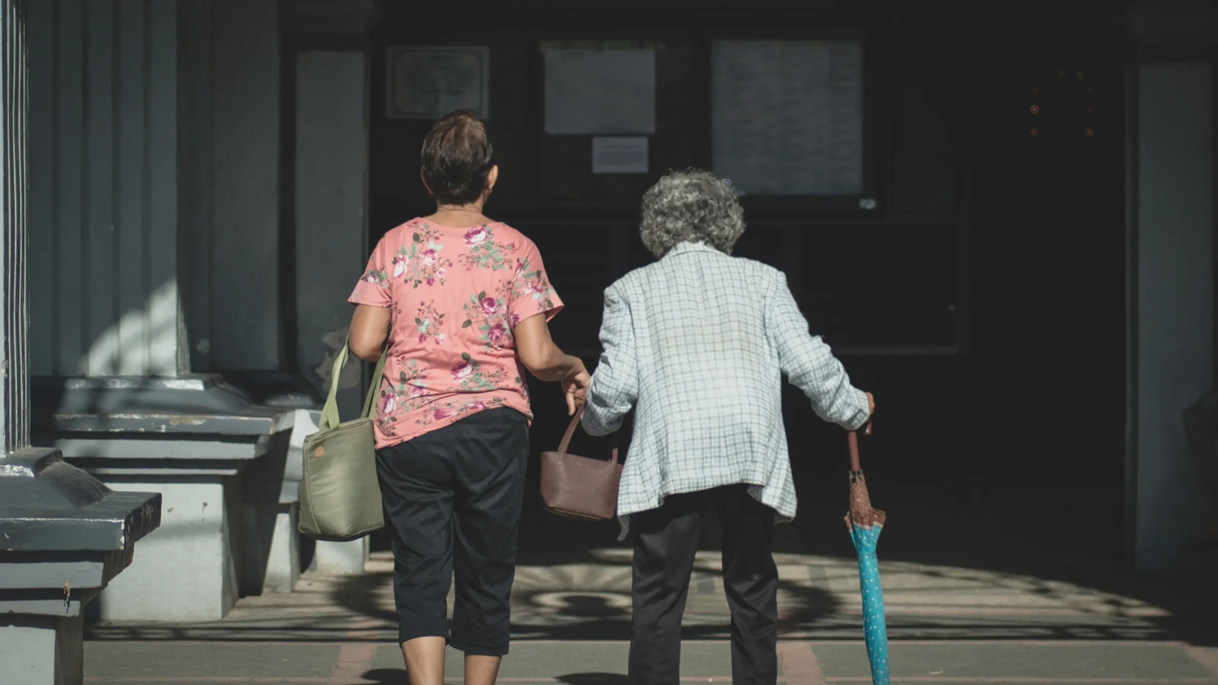 A woman walking close beside her elderly mother, carrying her bag as they head inside together