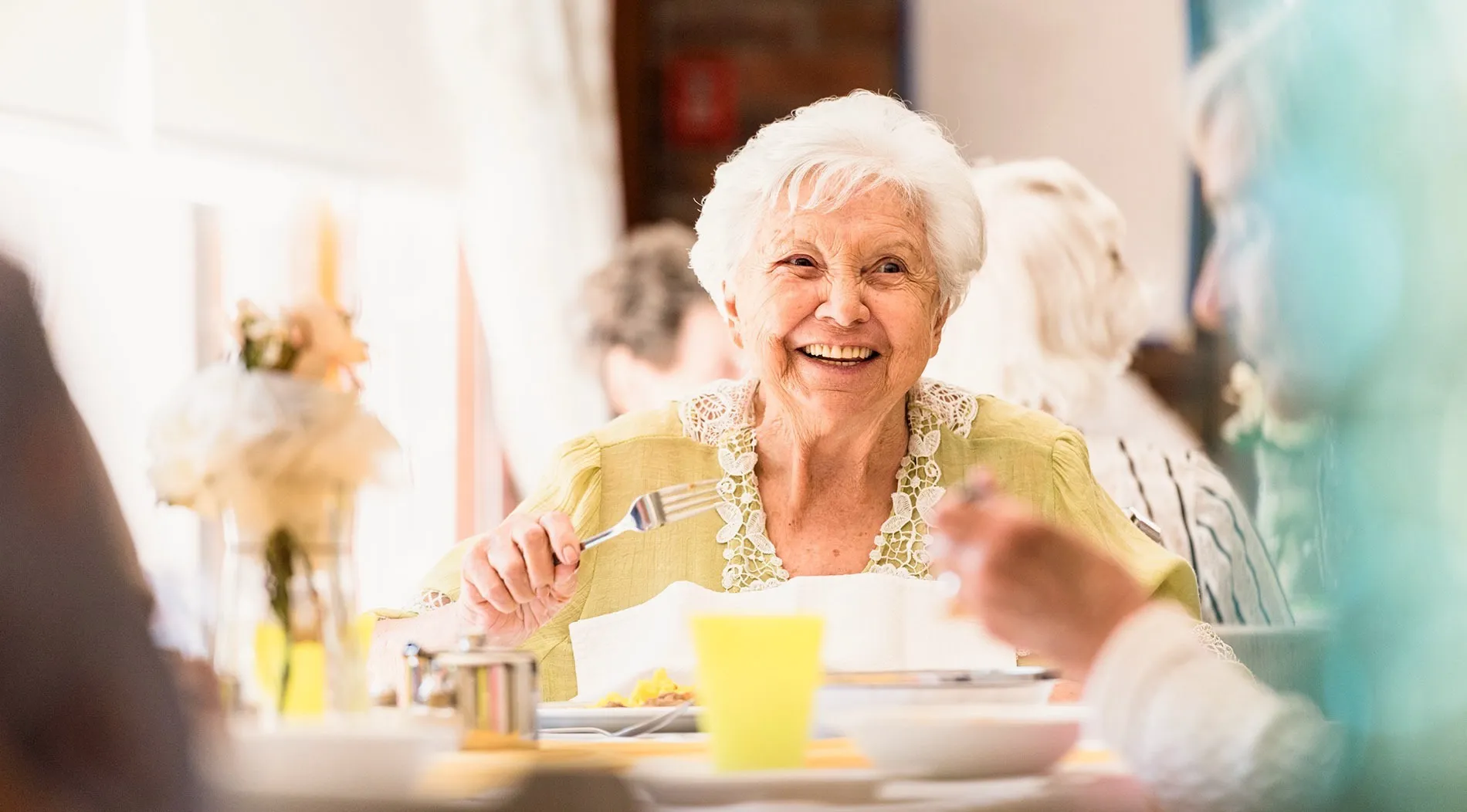 A smiling older woman enjoying a meal with friends in a sunny dining room